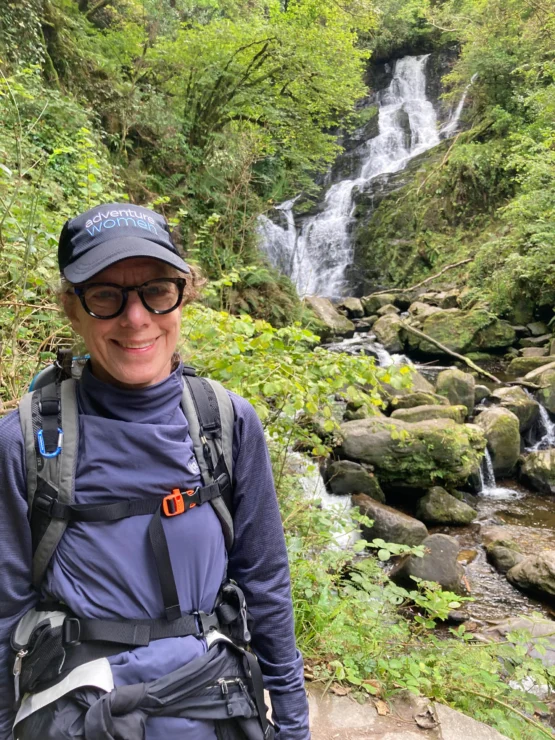 A person wearing a cap and glasses stands smiling in front of a waterfall surrounded by greenery.