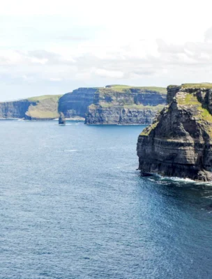 Cliffs meet the ocean under a cloudy sky, with dark rock faces and grassy tops extending into the distance. Waves gently crash against the cliff base.