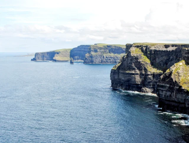 Cliffs meet the ocean under a cloudy sky, with dark rock faces and grassy tops extending into the distance. Waves gently crash against the cliff base.