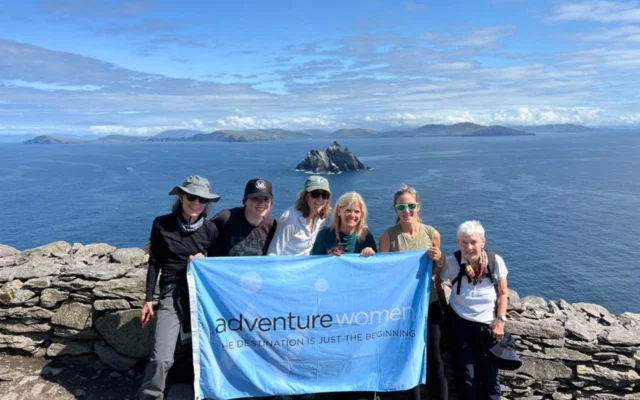 A group of women holding an "Adventure Women" banner stands on a rocky coastal viewpoint with islands in the background.