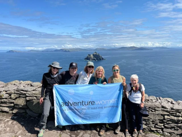 A group of women holding an "Adventure Women" banner stands on a rocky coastal viewpoint with islands in the background.