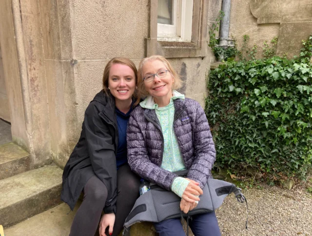 Two women sitting on stone steps outside, smiling at the camera. One is wearing a black jacket, and the other a gray jacket. Ivy grows on the stone wall beside them.
