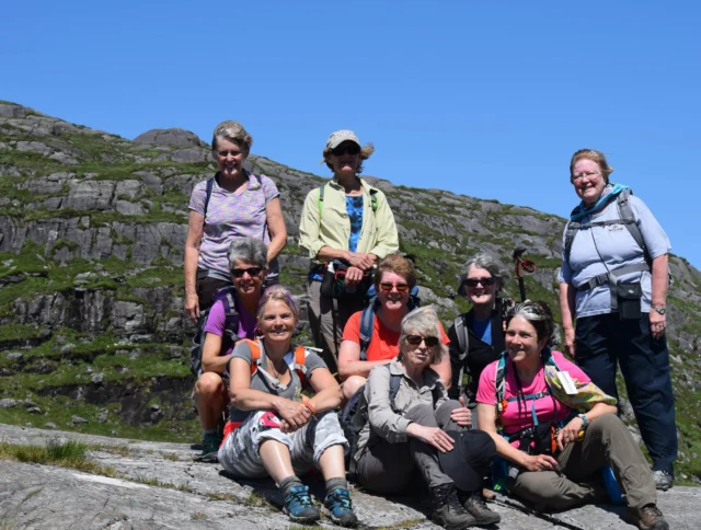 A group of nine people, mostly women, sitting and standing on rocky terrain with a clear blue sky in the background. They're dressed in outdoor gear, some with backpacks and hiking poles.