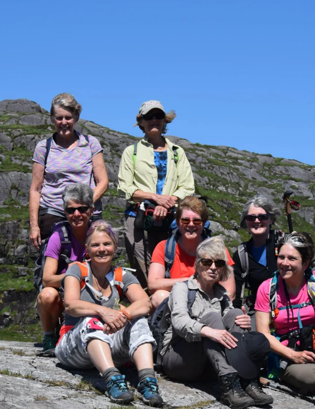 A group of nine people, mostly women, sitting and standing on rocky terrain with a clear blue sky in the background. They're dressed in outdoor gear, some with backpacks and hiking poles.