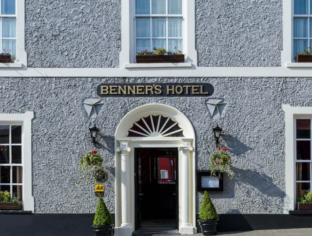 Front view of Benner’s Hotel with a gray exterior, white trim, and hanging plants flanking the entrance. Two upper-story windows have flower boxes.