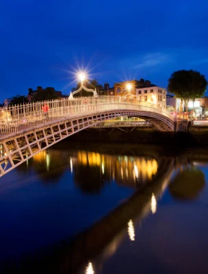 Illuminated arched pedestrian bridge over a calm river at night, with nearby buildings and streetlights reflecting on the water.