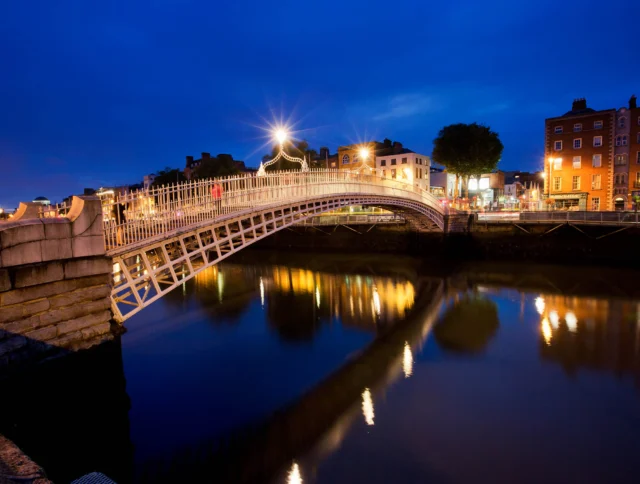 Illuminated arched pedestrian bridge over a calm river at night, with nearby buildings and streetlights reflecting on the water.