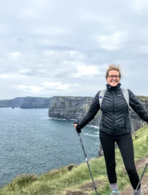 A person stands smiling on a coastal trail holding trekking poles, with cliffs and the sea in the background under a cloudy sky.