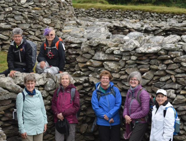 A group of seven people pose among stone ruins, wearing outdoor clothing and backpacks. Some are smiling; the background is grassy.