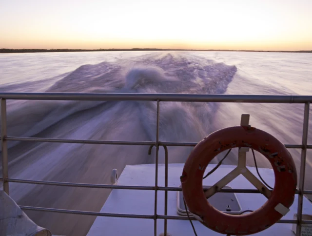 View from a boat's stern showing the wake trailing behind as it moves across a vast body of water at sunset. A red lifebuoy is attached to the railing.