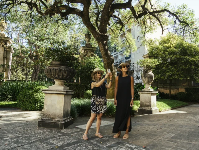 Two people in hats stand on a stone path in a garden, surrounded by trees and plants. One takes a photo with a smartphone.