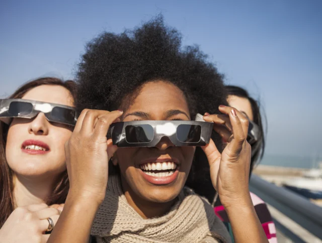 Three people smiling and looking up while wearing solar eclipse glasses outdoors.