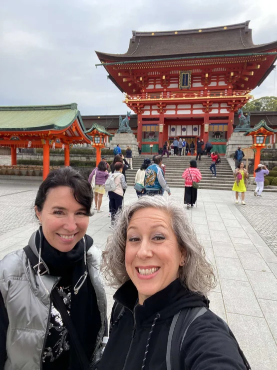 Two women smiling in front of a historic Japanese shrine with people walking around.