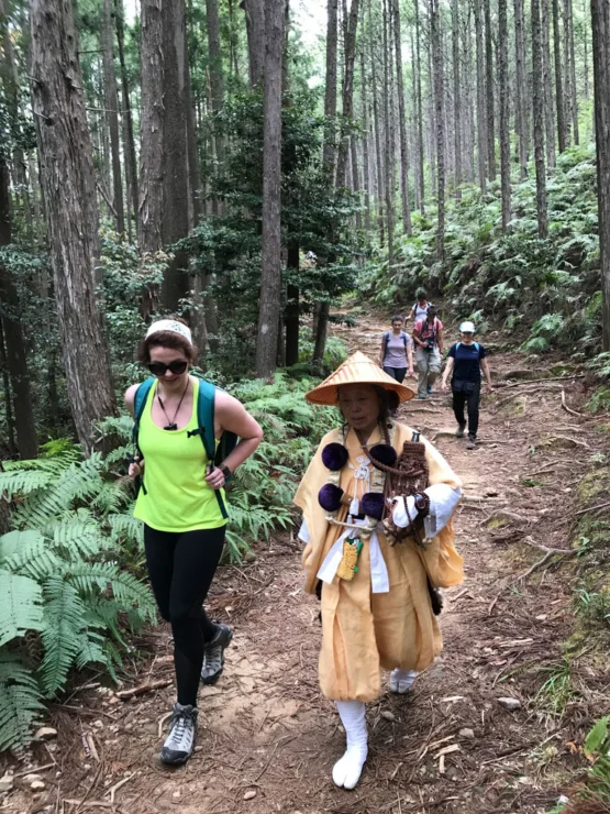 People hiking on a forest trail, including a person in traditional attire and others in casual hiking clothes, surrounded by tall trees and greenery.