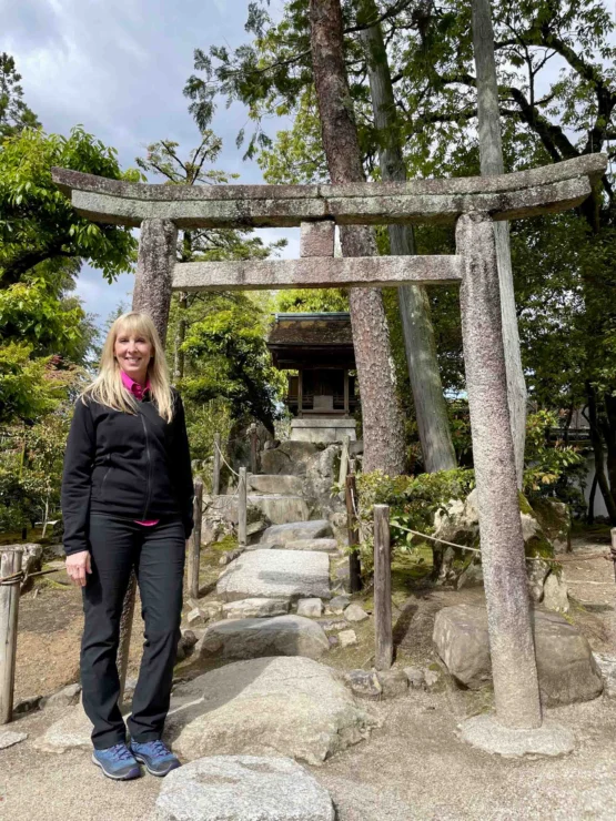 A person stands beside a traditional stone torii gate in a peaceful garden setting with greenery and a stone path.