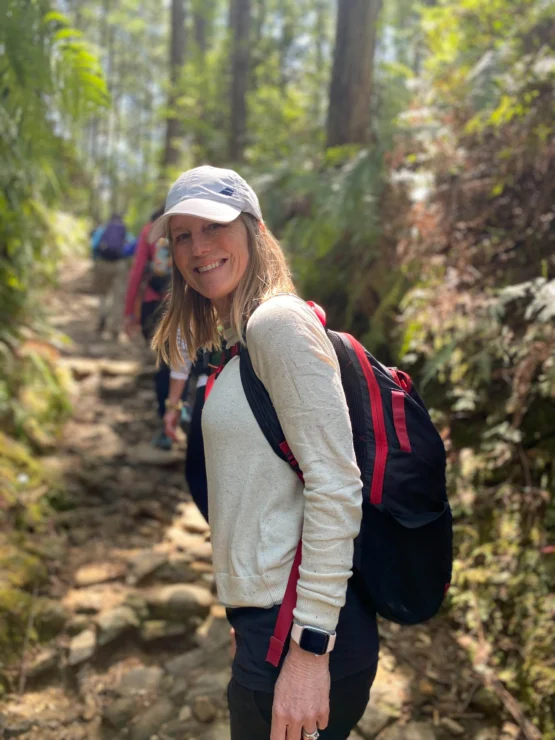 A woman in a white cap and sweatshirt hikes on a forest trail, smiling. She carries a black and red backpack.