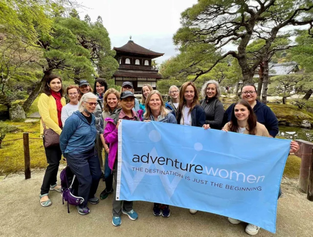 A group of women stand outdoors holding an "Adventure Women" banner, with a traditional Japanese building and trees in the background.