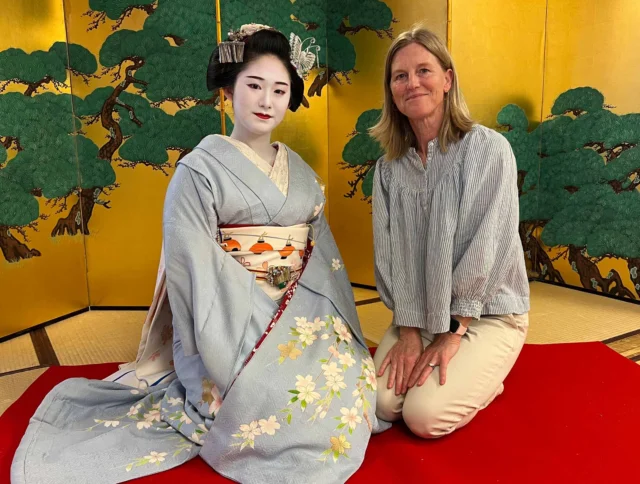 A woman in a kimono and another woman sit on a red mat in front of a decorative screen with tree motifs.