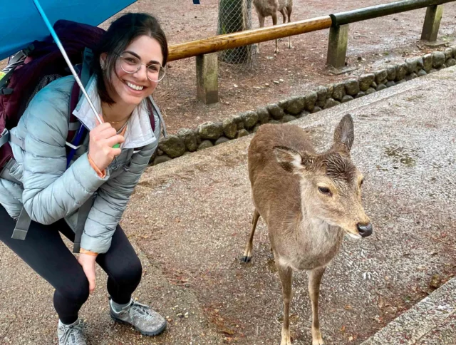 A person with glasses and an umbrella smiles next to a deer on a wet pathway.