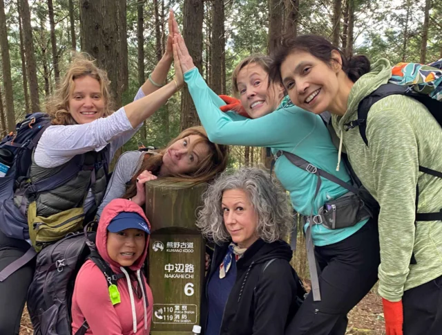 A group of six women in hiking gear pose cheerfully around a wooden signpost in a forested area.