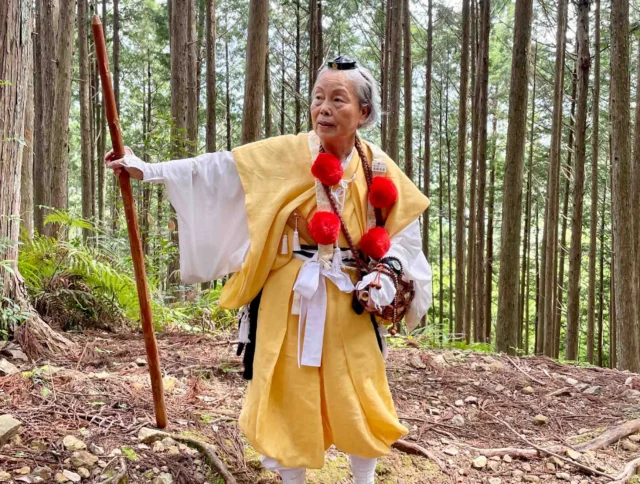 Elderly person in traditional yellow and white attire with red pom poms stands with a walking stick in a forest.