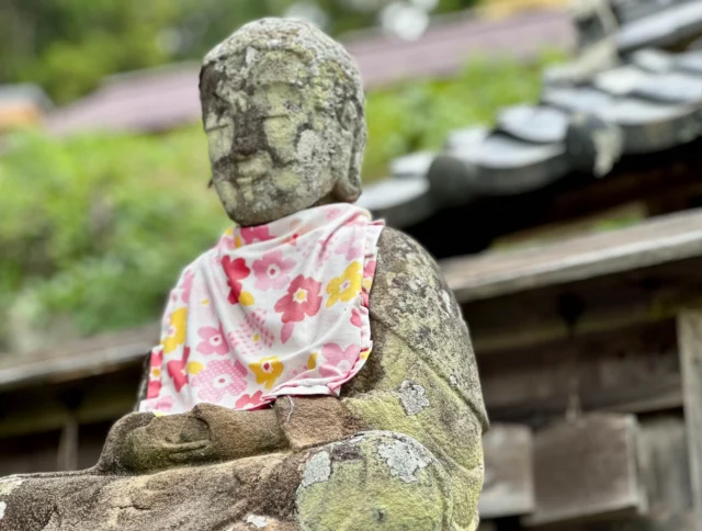 A weathered stone statue wearing a floral-patterned cloth sits outdoors, with a backdrop of greenery and wooden structures.