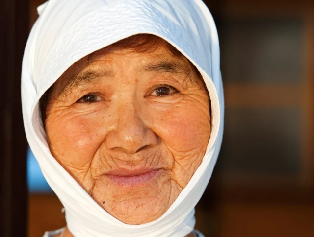 An elderly person wearing a white headscarf smiles softly, with a sunlit face, against a blurred outdoor background.