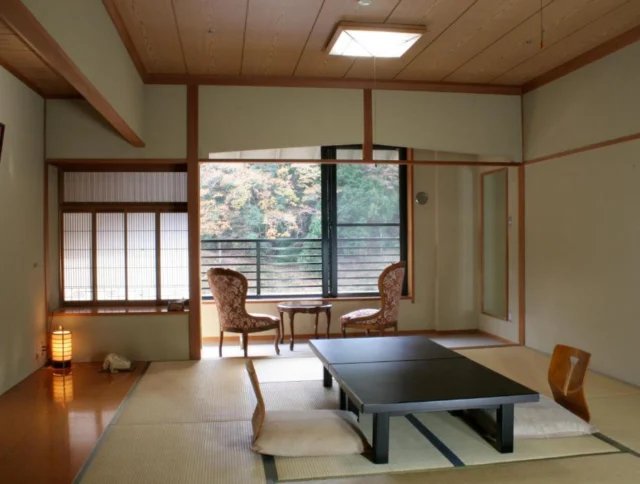 A Japanese-style room with tatami mats, a low table, cushions, and chairs by a window offering a view of nature.