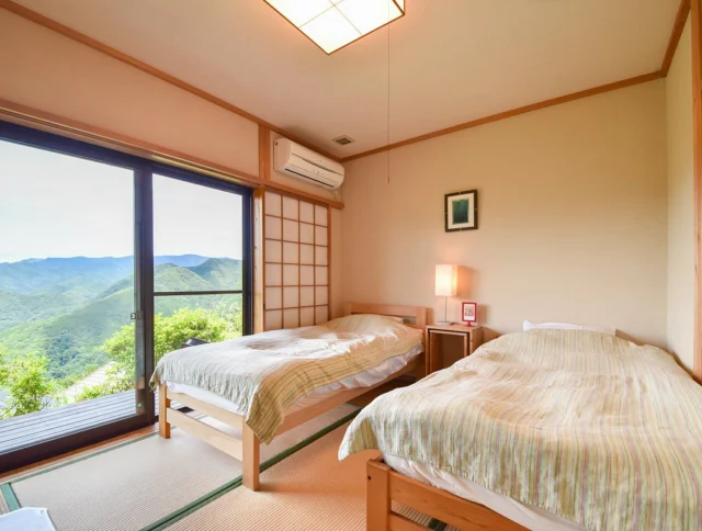 A serene bedroom with two beds, tatami mats, a sliding glass door leading to a balcony, a view of mountains, and a paper lantern ceiling light.