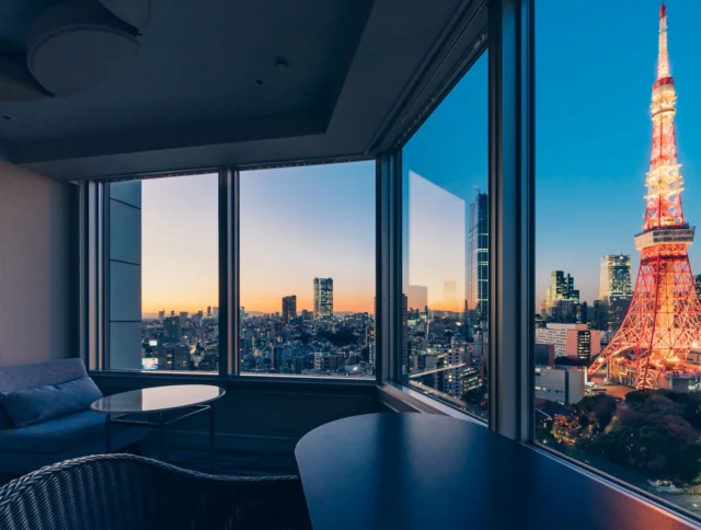 A hotel room with a view of a cityscape and a lit tower at dusk, visible through large windows.