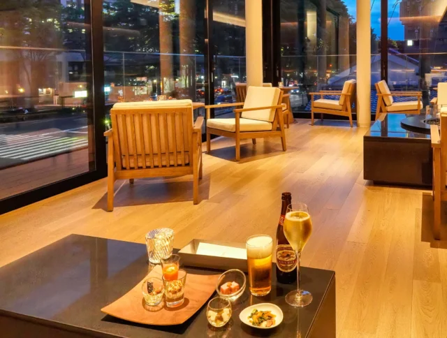 A modern lounge with wooden furniture overlooks a cityscape. A table in the foreground has drinks and appetizers, lit by candlelight. Large windows reveal the evening city lights outside.
