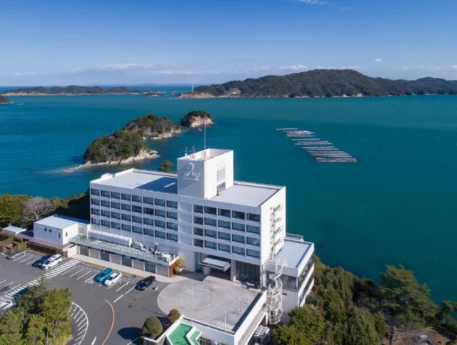 Aerial view of a white building by a blue ocean, with small islands and coastline in the background.