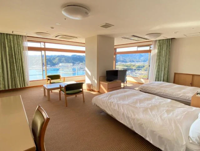 A hotel room with two beds, a desk, chairs, and a TV. Large windows reveal a scenic view of mountains and water.