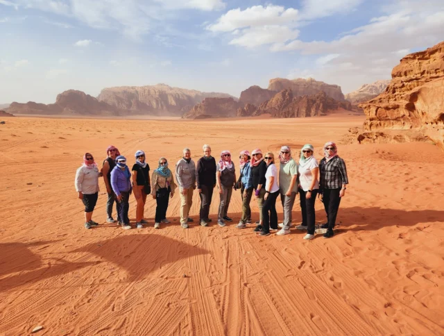 A group of people stands on a vast red desert landscape, with rock formations and a cloudy sky in the background.
