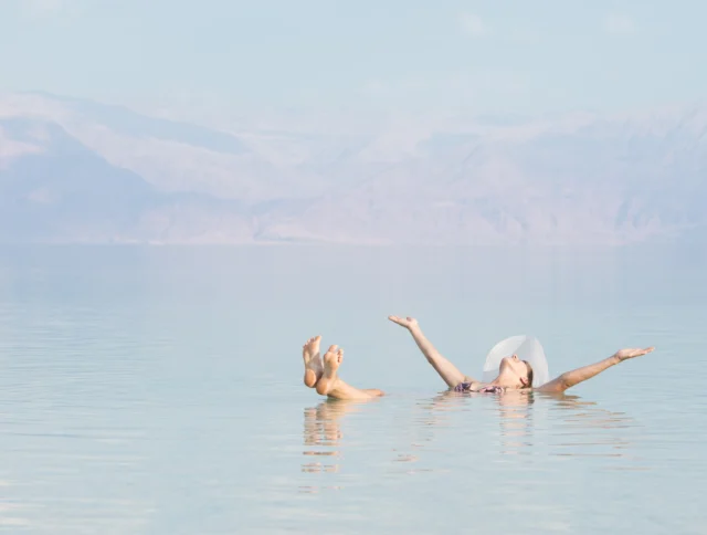 Person floating in a calm, clear body of water with mountains in the background, arms stretched out and wearing a white hat.