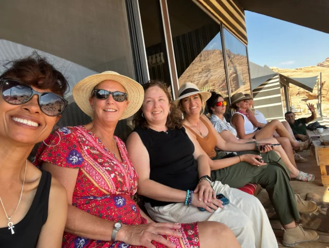 A group of people sitting outdoors on a wooden bench, wearing casual summer clothing and hats, with a desert landscape in the background.