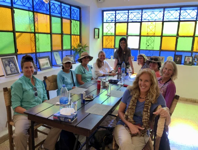 A group of nine people seated around a table with drinks and snacks in a room with colorful stained glass windows.