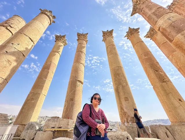 A woman stands in front of ancient stone columns under a clear blue sky. Another person is seen leaning against a column in the background.