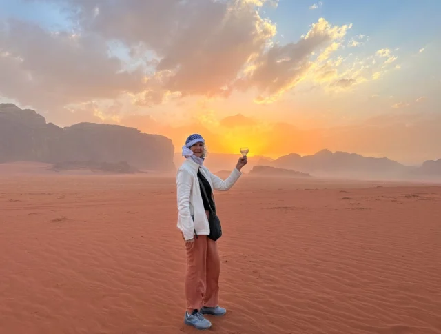 Person standing in a desert at sunset, holding a glass, wearing a white jacket and headscarf. Rocky formations and clouds are visible in the background.