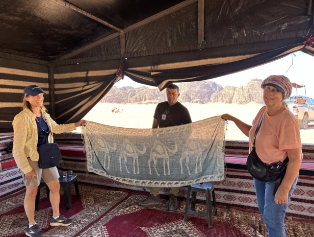Three people hold a camel-patterned mat inside a tent with desert scenery in the background.