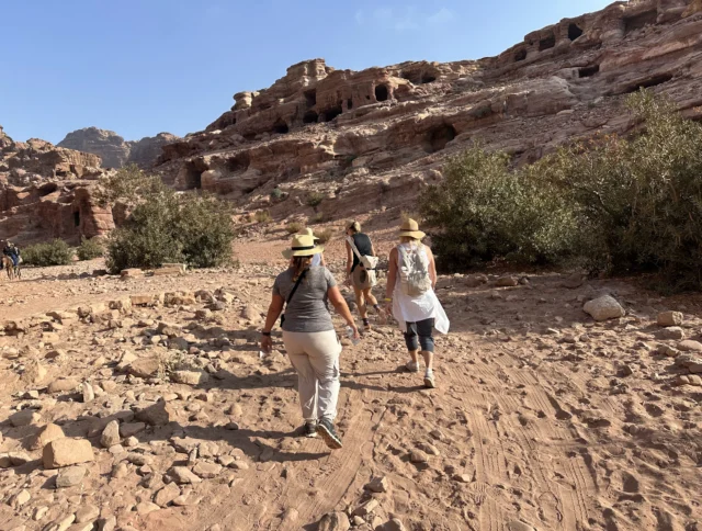 Three people wearing hats walk along a rocky path in a desert landscape with cliffs and greenery under a clear blue sky.