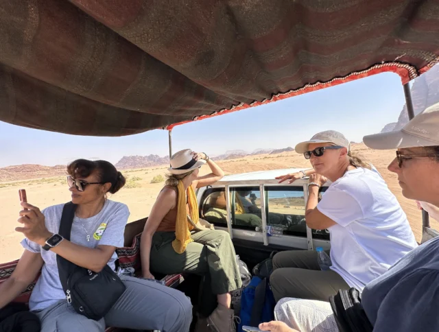 Four people sitting in an off-road vehicle traveling through a desert landscape, one taking a photo, others looking at the scenery.