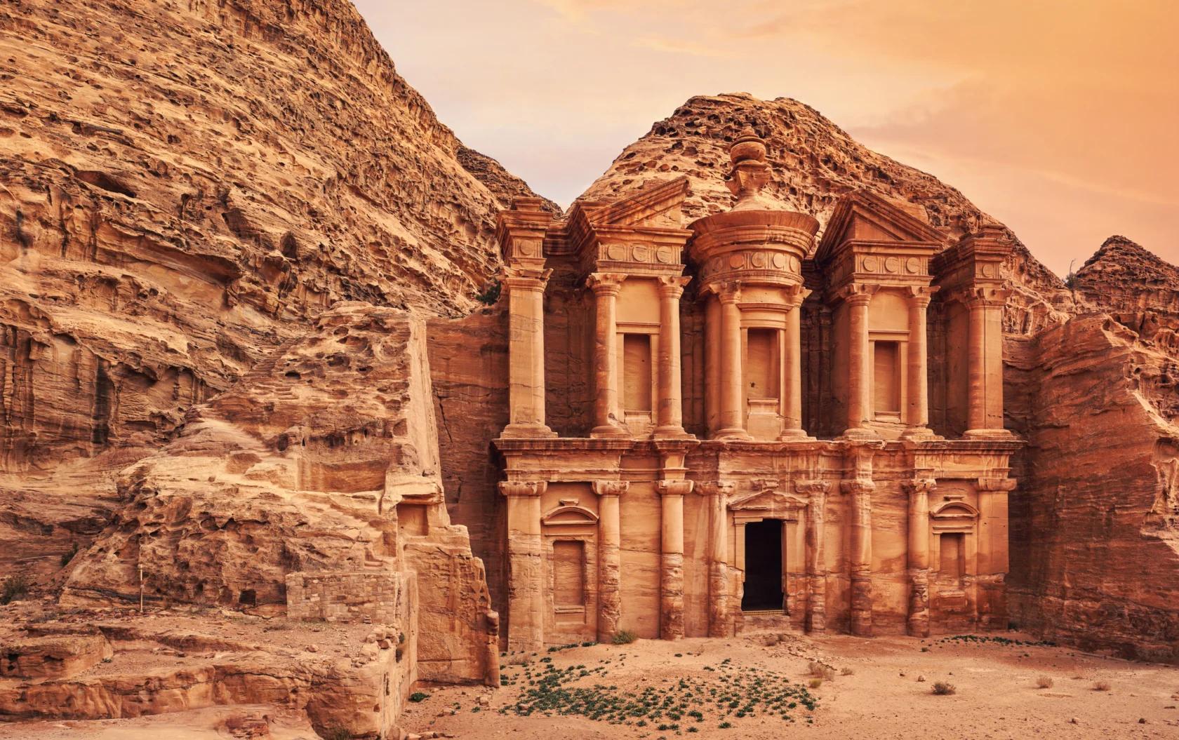The image shows the ancient rock-cut architecture of Al Deir, the Monastery, in Petra, Jordan, with a mountainous backdrop and an orange sky.