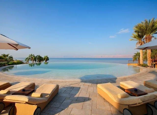 Infinity pool with lounge chairs overlooking a vast body of water and palm trees under clear blue skies.
