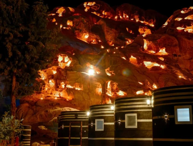 Cave lit up at night with warm lights highlighting its contours; striped, circular huts with windows are in the foreground; a tree stands to the left.