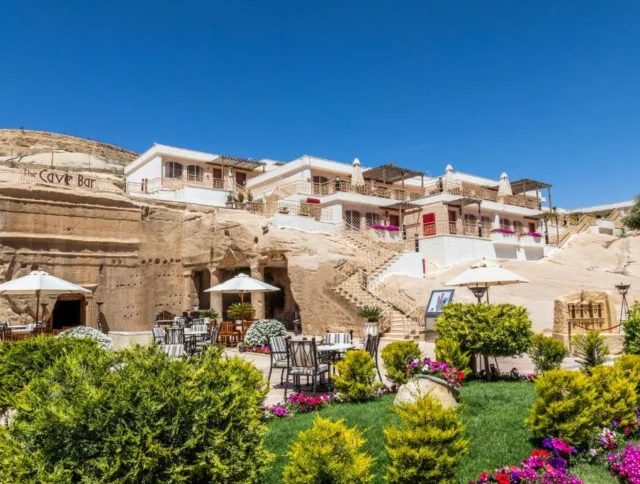 A hillside hotel with stone buildings, outdoor seating, and green landscaping under a clear blue sky.