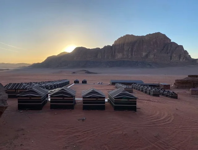Sun setting over a desert landscape with rows of striped tents and rocky mountains in the background.