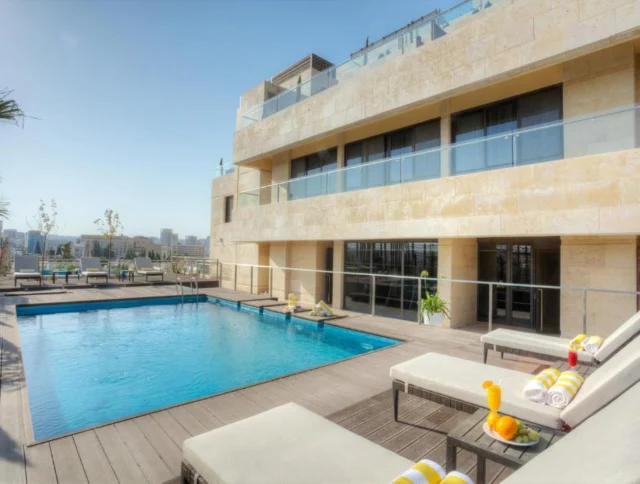 A modern hotel rooftop with a swimming pool, sun loungers with towels, and city skyline in the background on a clear day.