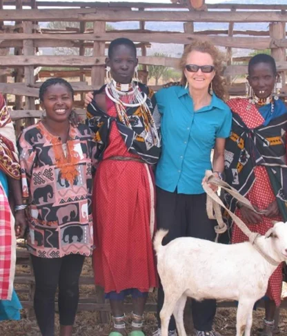 A group of people, including a woman in a blue shirt, stand together outdoors with a white goat. They are wearing colorful clothing and smiling. A wooden fence is in the background.
