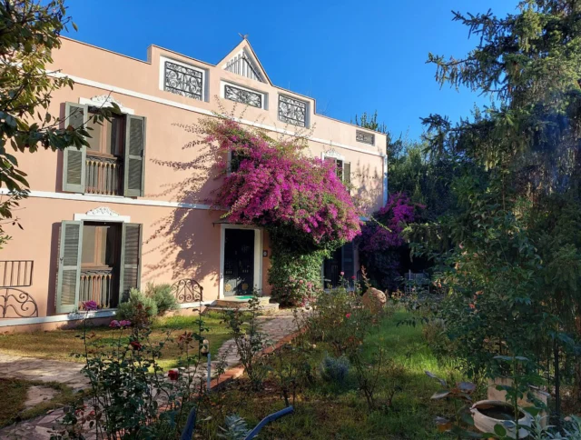 A two-story peach house with purple bougainvillea climbing the front. Surrounded by a garden with lush greenery and a clear blue sky above.
